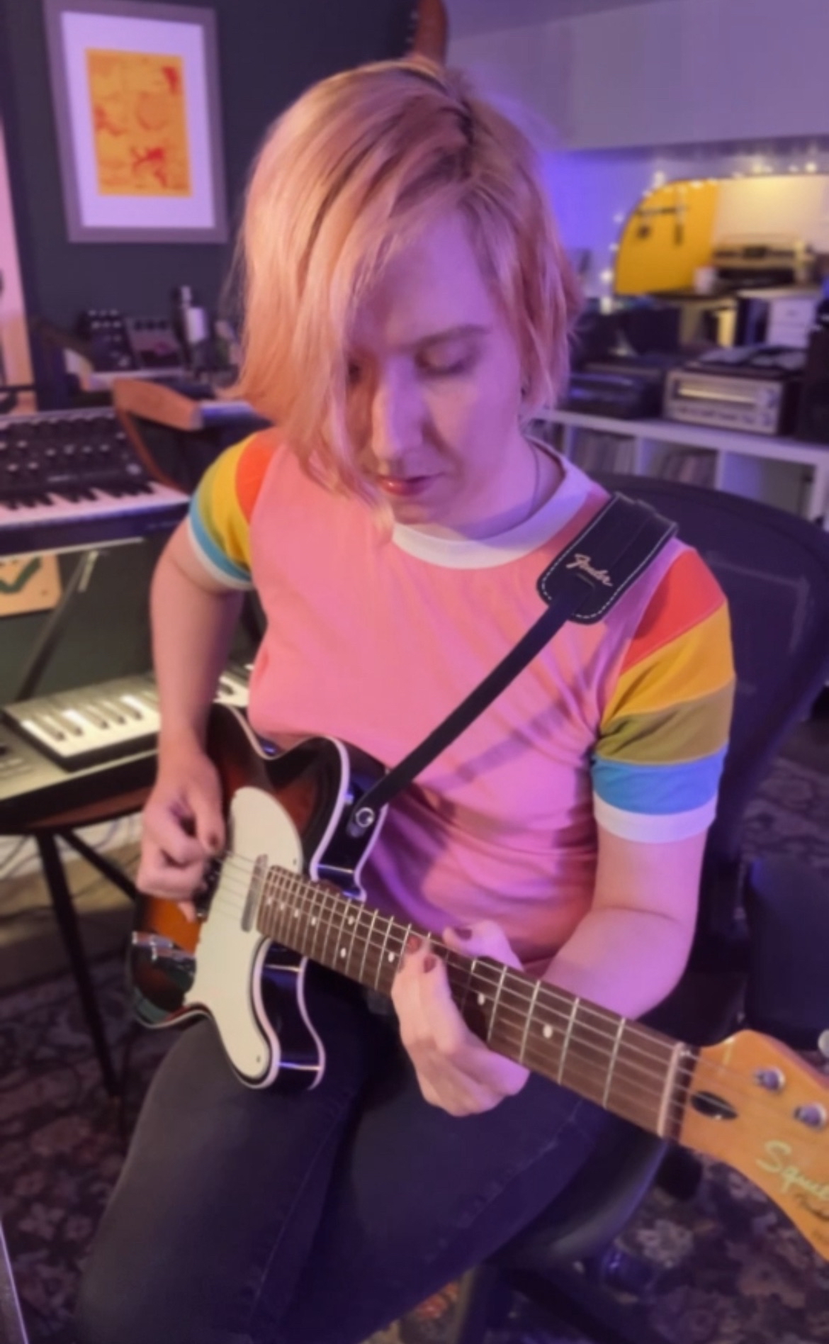 A blonde-haired girl in a pink shirt with rainbow sleeves playing a squier telecaster in sunburst. The room is lit with violet light.