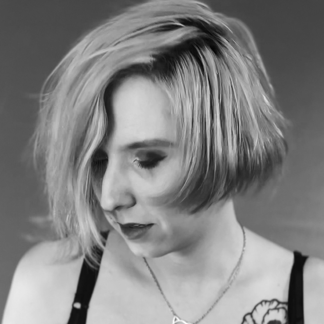 Lex Feathers, a woman with light hair and dark makeup. She has her eyes closed and head tilted downward.